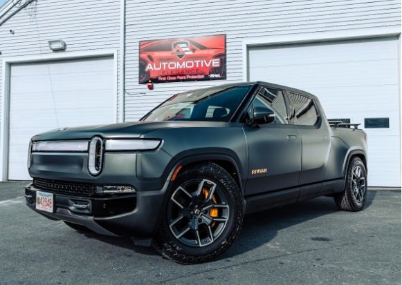 A dark matte grey Rivian R1T truck parked outside a garage with a red 'Automotive Elegance' sign.