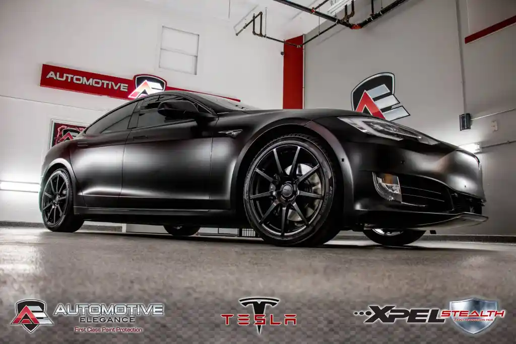 Matte black Tesla Model S with black rims, parked in a garage with Automotive and Tesla logos.
