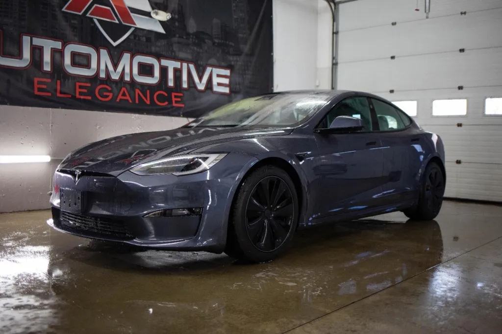 A dark grey Tesla Model S covered in water droplets, parked on a wet garage floor, in front of a banner.