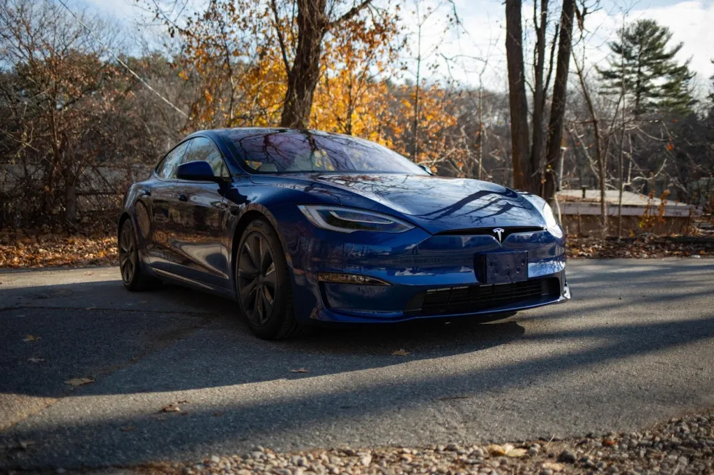 A highly reflective dark blue Tesla Model S parked on an asphalt road with autumn trees in the background.