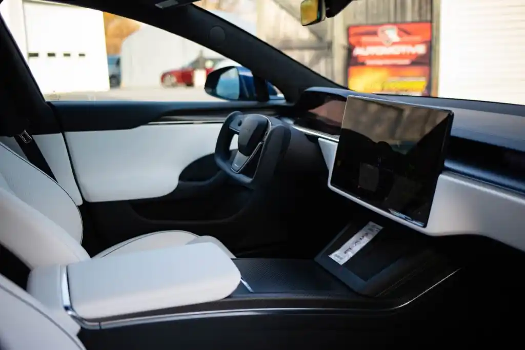 Driver's perspective inside a modern car with white and black interior, yoke steering wheel, and large central screen.