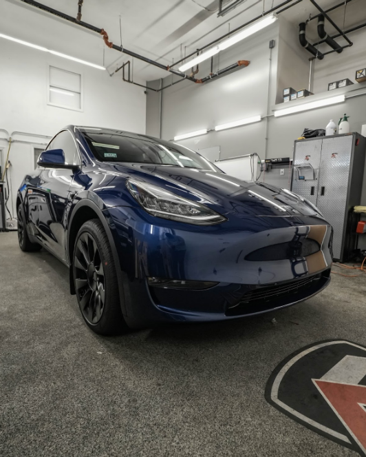 A dark blue Tesla Model Y, seen from a low angle, parked in a well-lit garage with industrial fixtures.