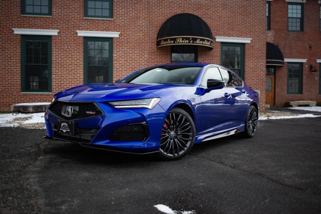 A sleek, bright blue Acura TLX Type S parked on an asphalt lot in front of a historic brick building.