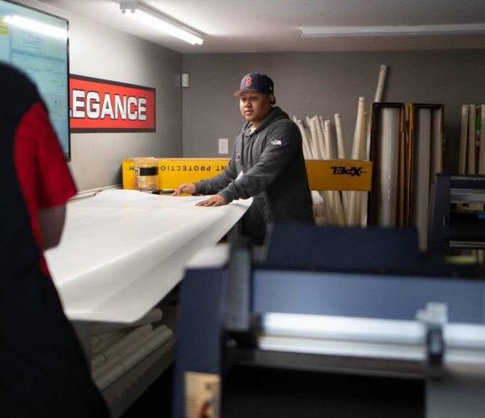 A man in a baseball cap works with large sheets of white material in a workshop, another person's back is in the foreground.