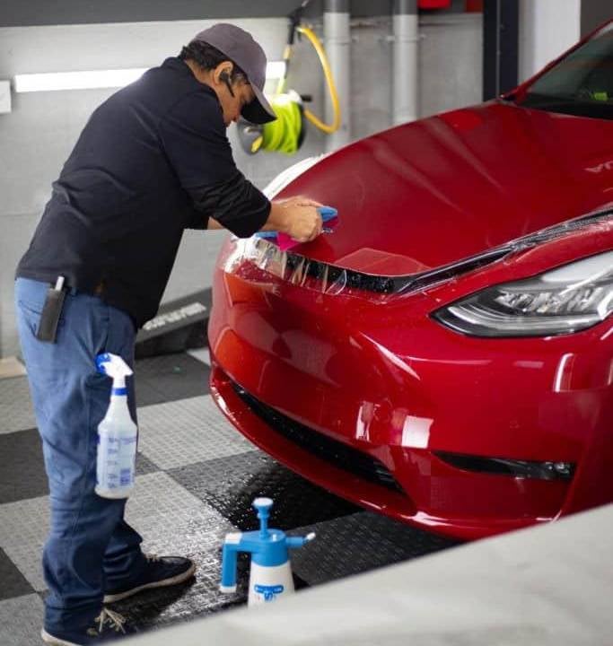 Person in a cap and dark shirt applying a clear protective film to the hood of a red Tesla car.