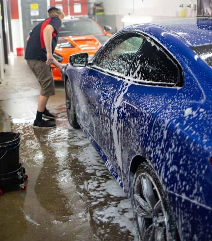 A blue car covered in white soap suds is being washed by a person in a garage with an orange car in the background.